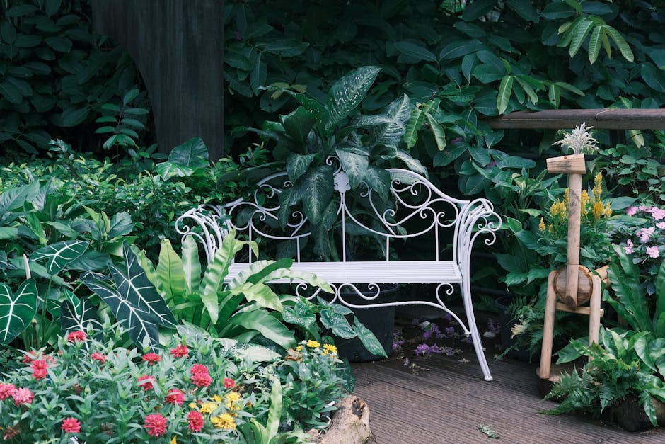 A lush garden scene featuring a white wrought iron bench positioned on a wooden deck, surrounded by a variety of vibrant plants and foliage. To the left, there are large, dark green leaves with promin