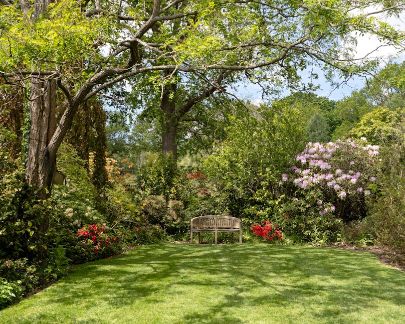 A lush garden scene featuring a white wrought iron bench positioned on a wooden deck, surrounded by a variety of vibrant plants and foliage. To the left, there are large, dark green leaves with promin