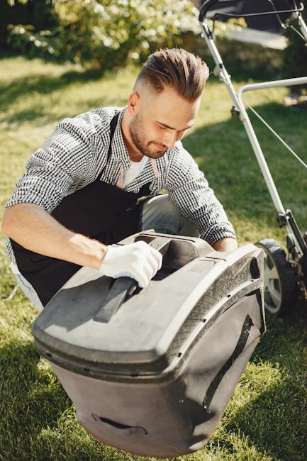 A man with neatly styled brown hair and a trimmed beard is engaged in garden maintenance outdoors under bright sunlight, kneeling on a well-maintained lawn. He is wearing a checkered shirt, black apro