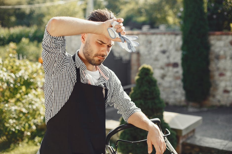A man with neatly styled brown hair and a trimmed beard is engaged in garden maintenance outdoors under bright sunlight, kneeling on a well-maintained lawn. He is wearing a checkered shirt, black apro